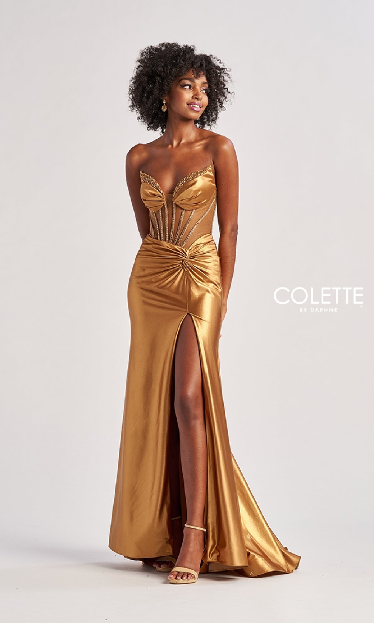 A woman wears the Sheer-Bodice Long Prom Dress: Colette CL8560 in metallic gold, featuring a strapless corset bodice, sheer paneling, gathered waist details, and a thigh-high slit, standing against a plain background.